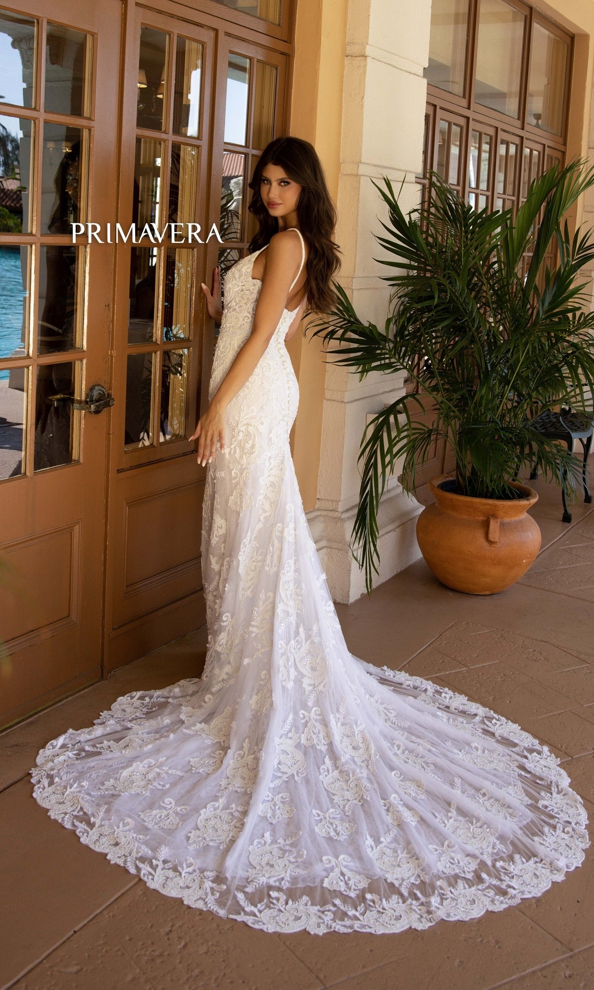 A woman in the Long Wedding Dress 11112 by Primavera, featuring ivory lace and a train, stands by glass doors and a potted plant, looking over her shoulder.